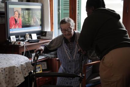 Ayuda. Una anciana se incorpora apoyada en un caminador y en los brazos de su cuidadora. No solo la edad sino también enfermedades y accidentes pueden hacer necesaria este tipo de asistencia a domicilio.